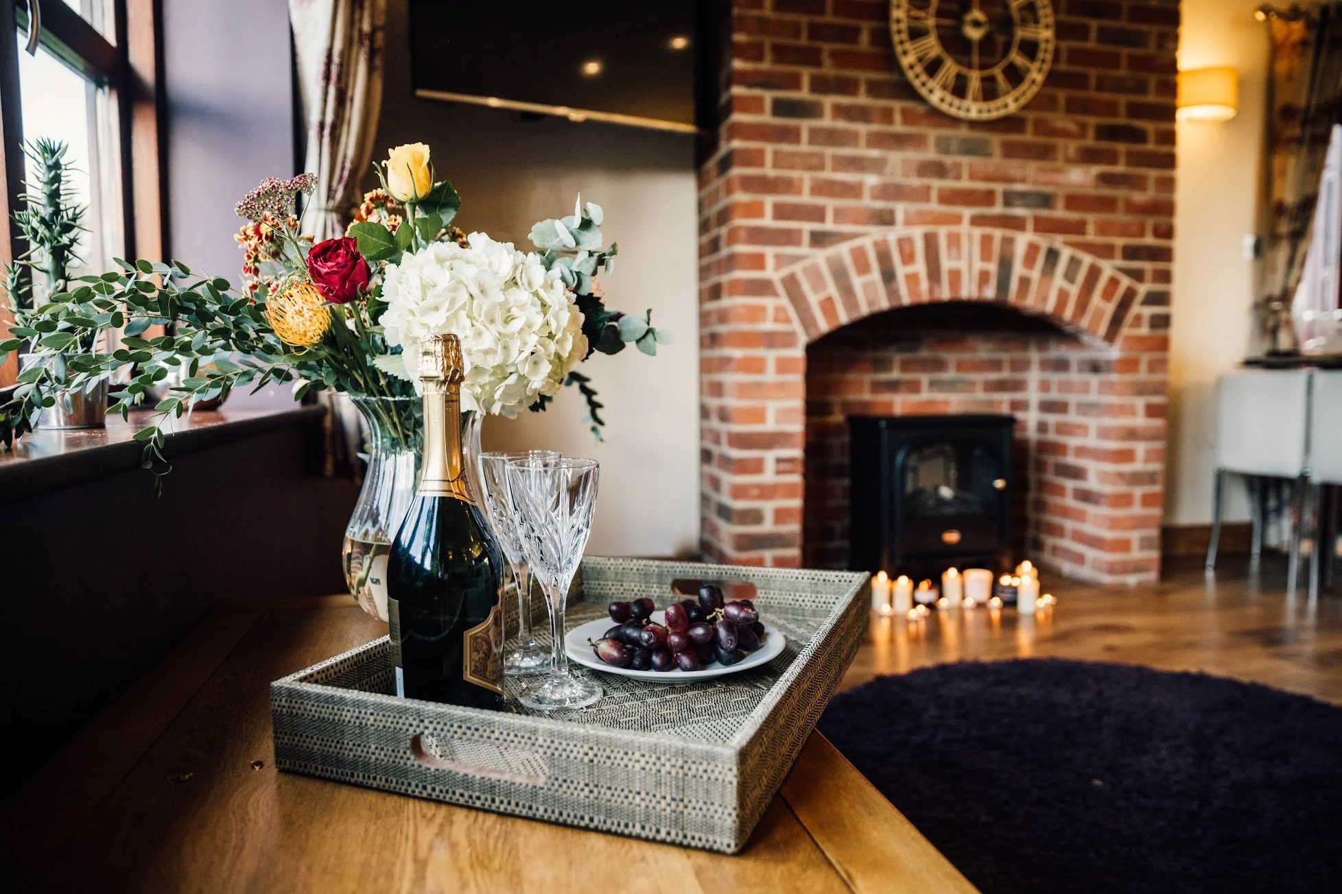 A table set with a bouquet of flowers in a glass vase, a bottle of champagne, two crystal wine glasses, and a plate of grapes in a cozy living room with a brick fireplace, candles on the floor, and a large clock on the wall.