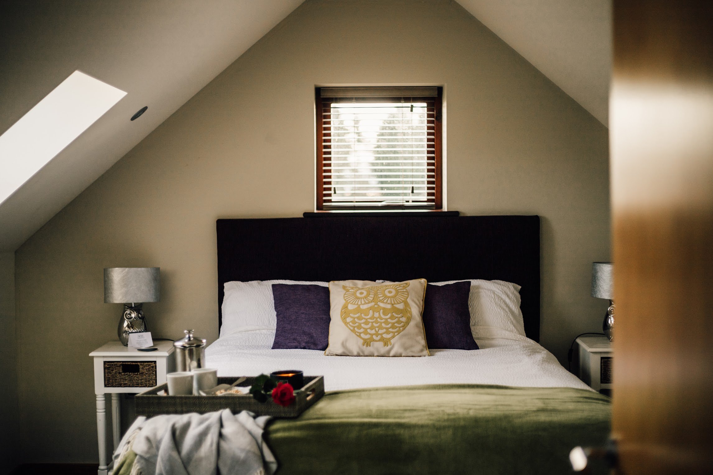 A bedroom with a sloped ceiling, featuring a window with blinds above the bed. The bed has pillows, including one with a gold owl design. There are two bedside tables with lamps on either side of the bed and a tray with cups and flowers on the foreground.
