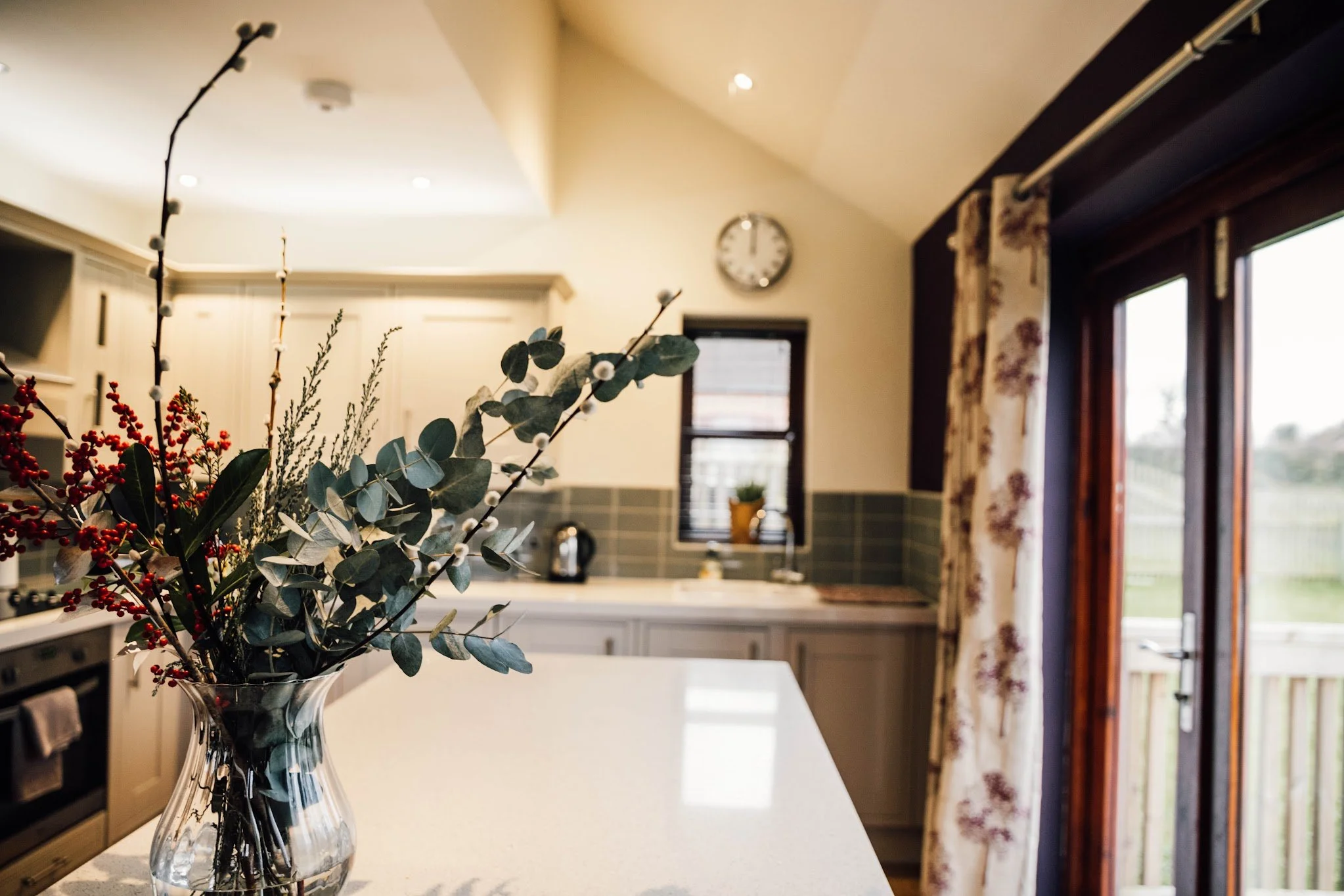 A clear glass vase with a bouquet of eucalyptus leaves, red berries, and other greenery on a kitchen countertop. In the background, there are windows with floral curtains, a clock on the wall, and a kitchen with white cabinets and gray backsplash.