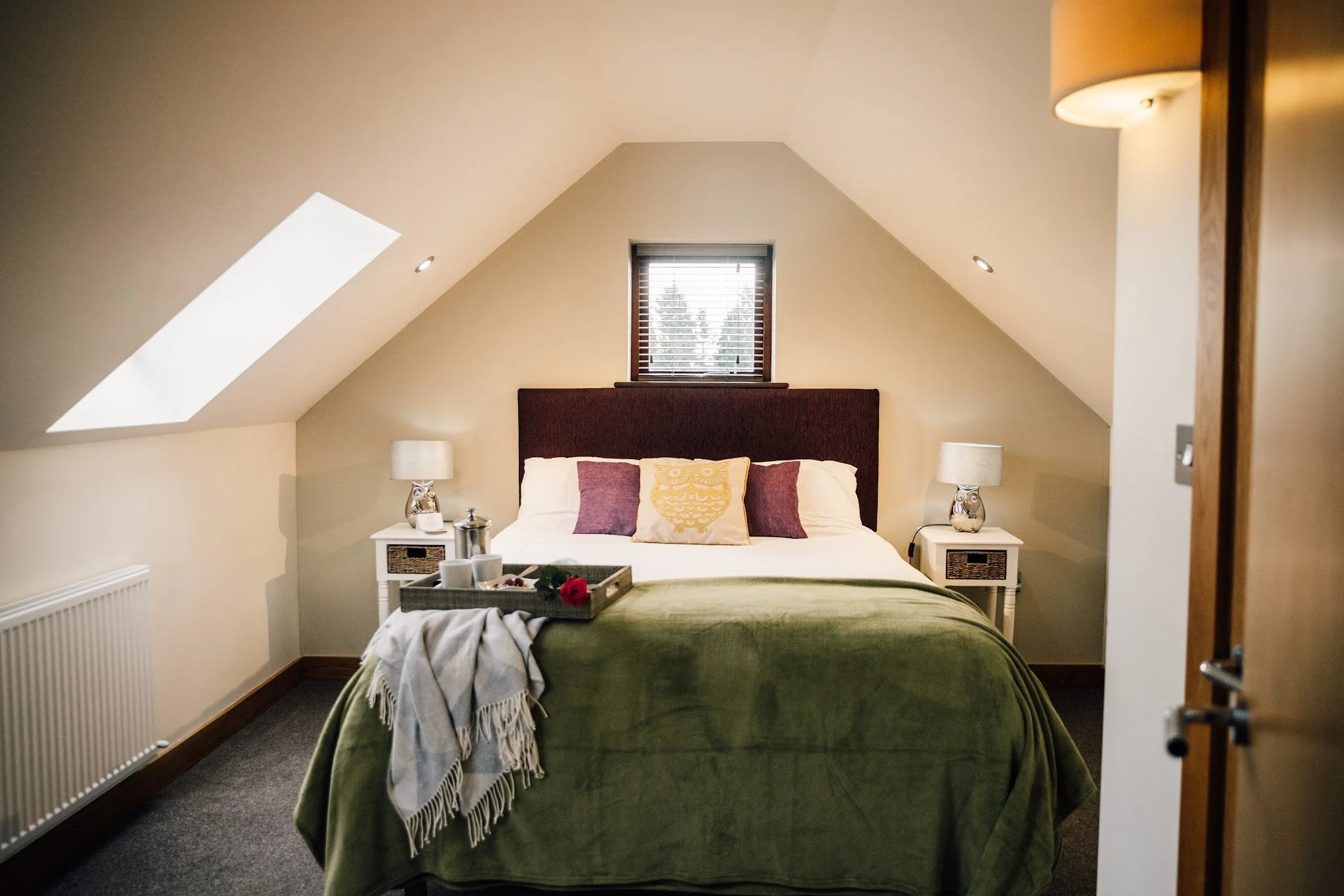 A cozy bedroom with a sloped ceiling, featuring a bed with a maroon headboard, four pillows including one with a yellow owl design, and a green bedspread. There are matching white nightstands with lamps on each side of the bed. A small window with bl