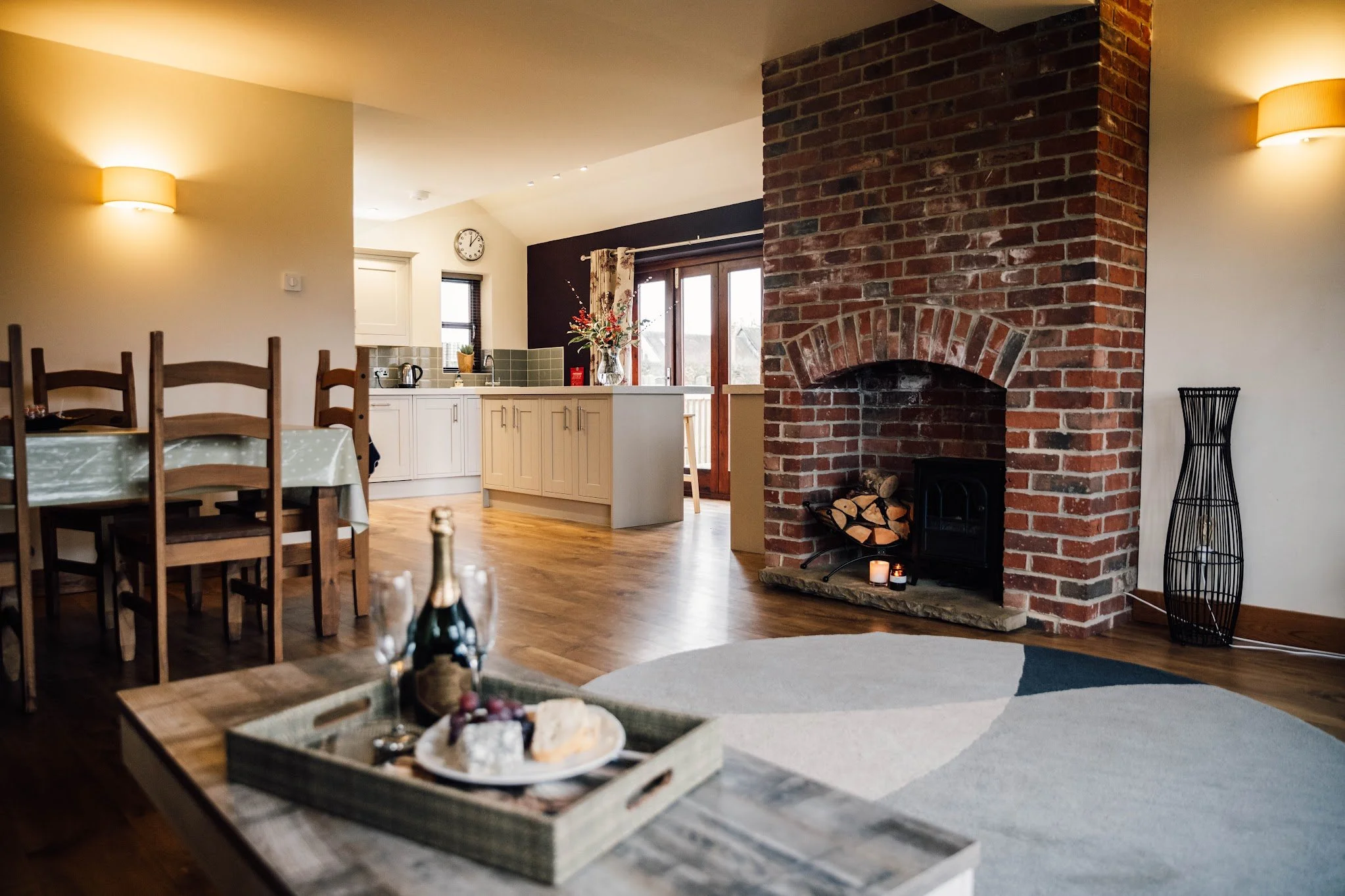 Open-plan living area with a brick fireplace, wooden dining table with chairs, kitchen with white cabinets, and sliding glass doors leading to a balcony, in a cozy home setting.