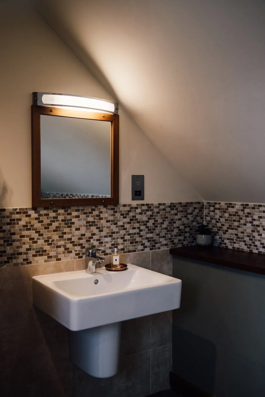 A small bathroom with a white sink, mirror with wooden frame, mosaic tile backsplash, wall-mounted light, and a dark wood surface on the right.