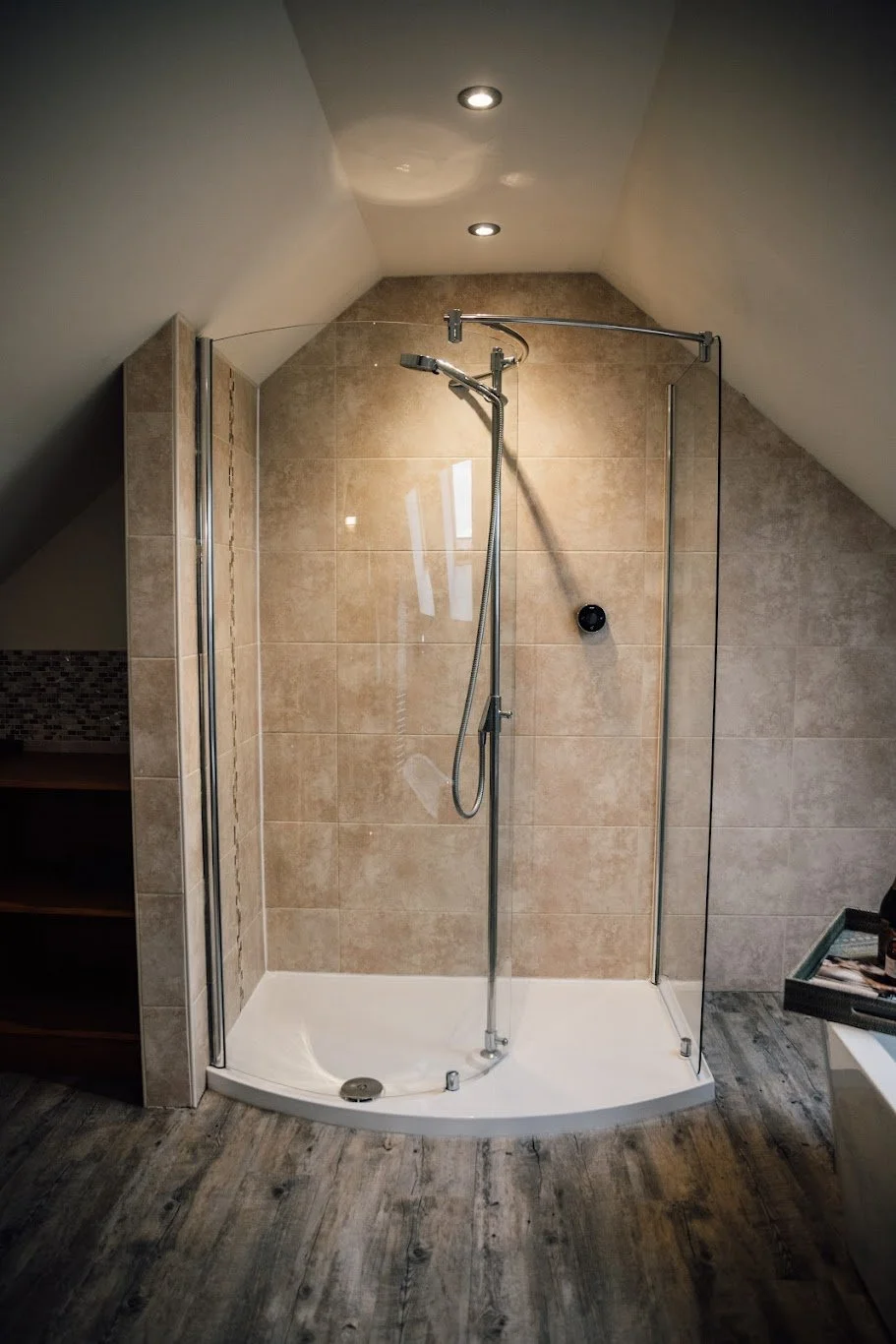 Empty corner walk-in shower with beige tiles, a handheld showerhead, glass enclosure, and ceiling lights in a bathroom with wood-look flooring.