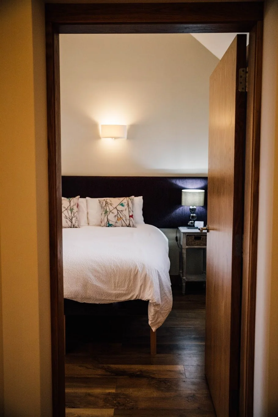 View into a bedroom through an open door, showing a bed with white bedding and decorative pillows, a nightstand with a lamp, and a wall-mounted light fixture.