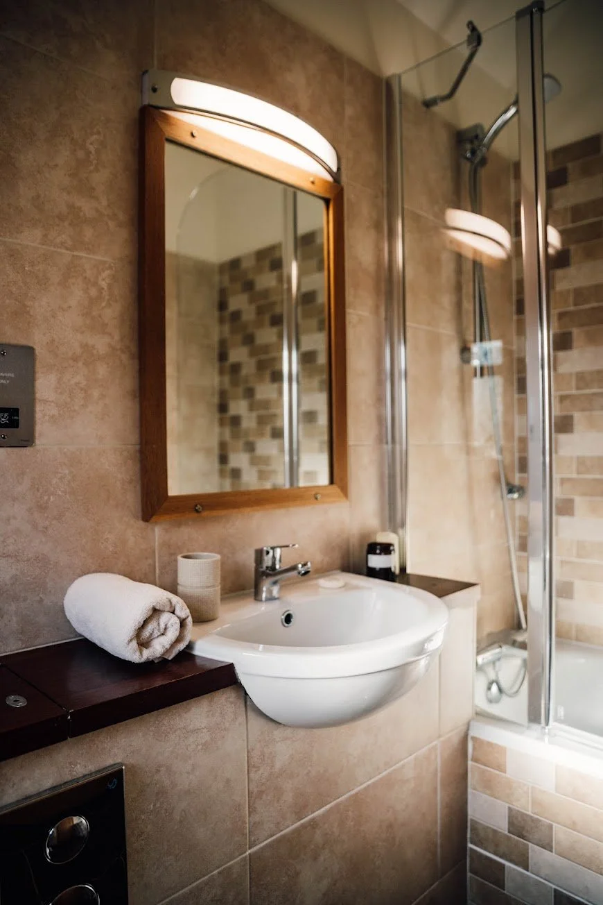 Bathroom with beige tiled walls, a small white sink with a mounted wooden mirror above, a rolled white towel, a toilet paper holder, and a shower with a glass door and brick-patterned wall tiles.