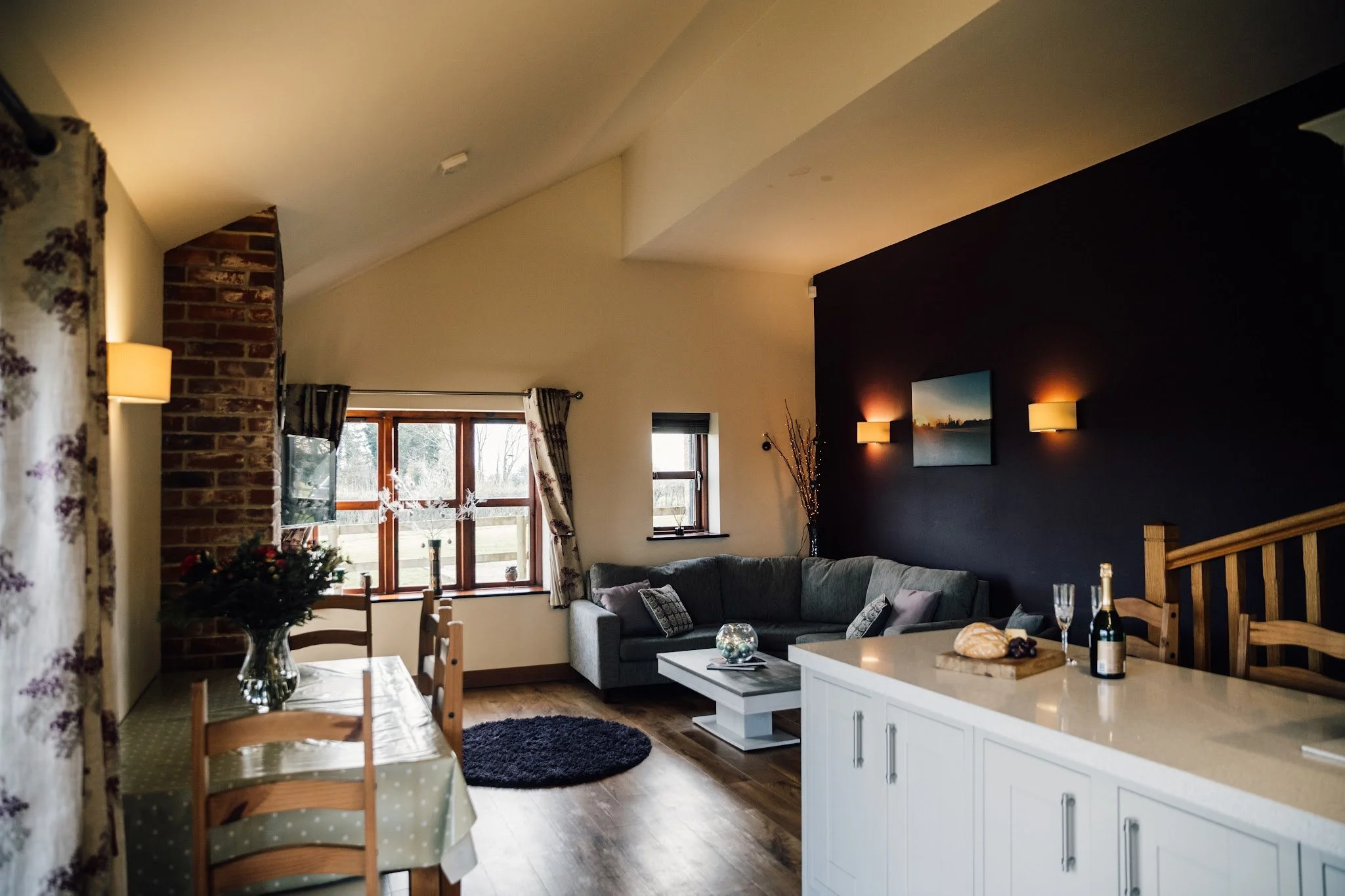 Living room with large windows, brick and dark-colored walls, a gray sofa with decorative pillows, a white coffee table with decorations, and a dining table with chairs. There are wall-mounted lights and framed pictures, with a view of trees outside.