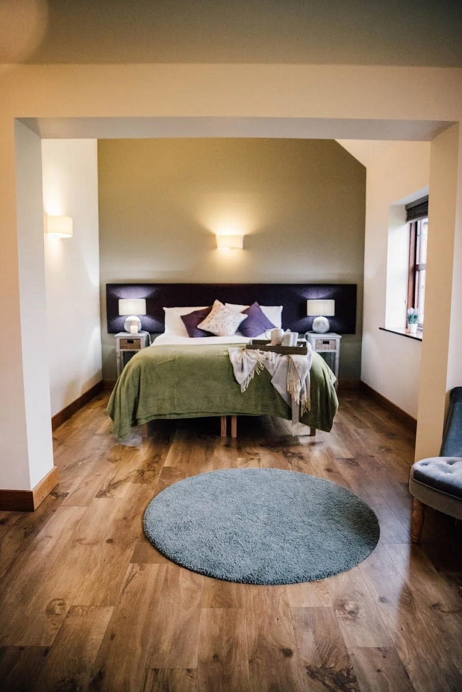 A cozy bedroom with a bed dressed in green and purple pillows, bedside lamps, a gray rug, wooden flooring, and a window with a small potted plant on the sill.