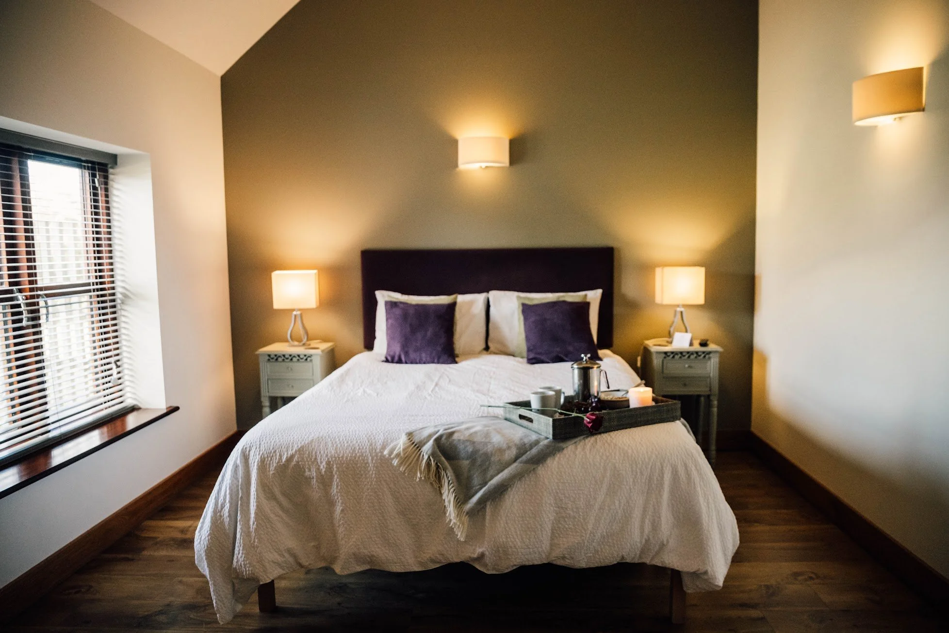 A cozy bedroom with a bed, purple and cream pillows, nightstands with lamps, wooden floors, and a tray of breakfast items on the bed.