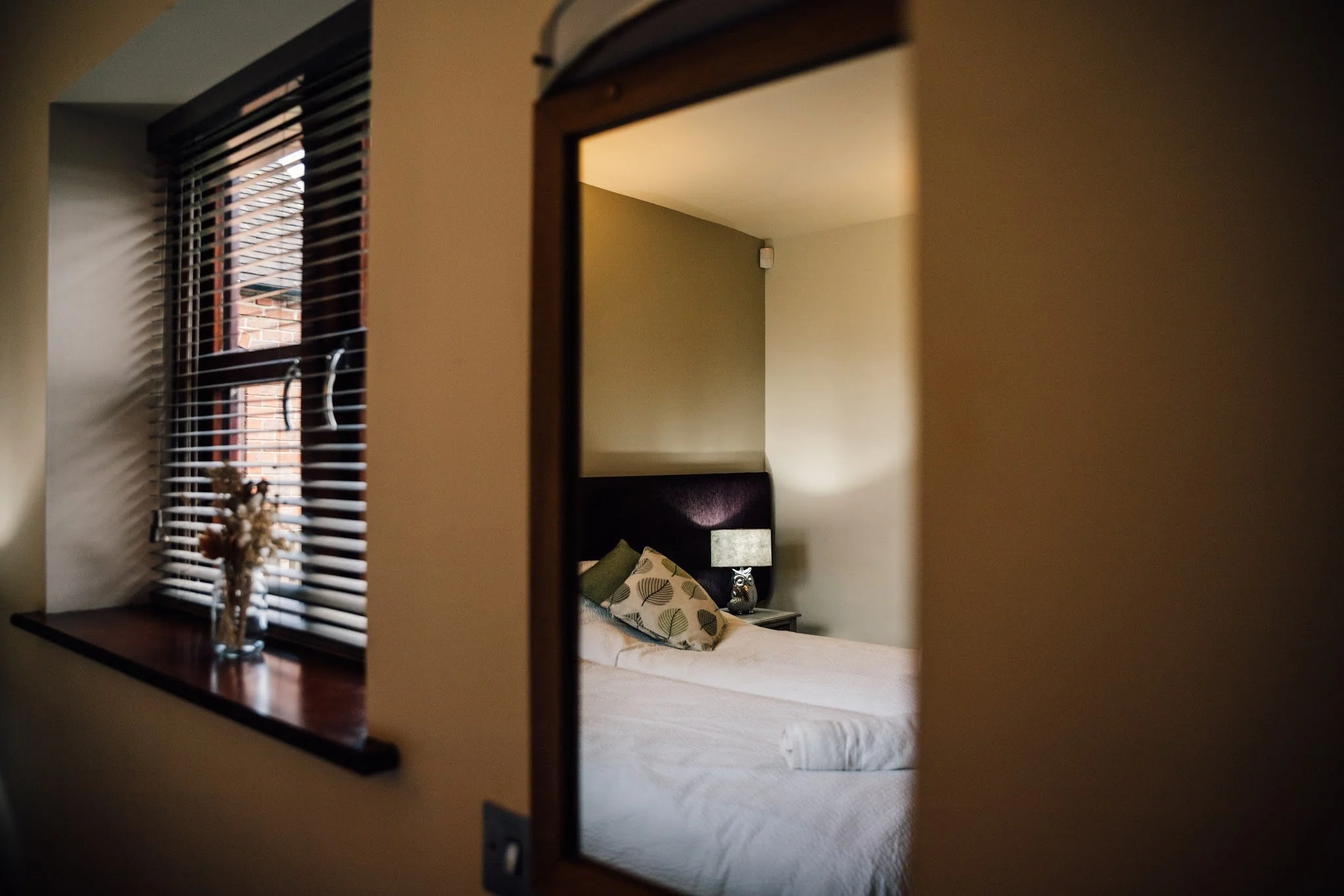 A mirror reflects a bedroom with a bed, pillows, and a nightstand with a owl figurine. There's a window with blinds and a small table with dried flowers outside the window.