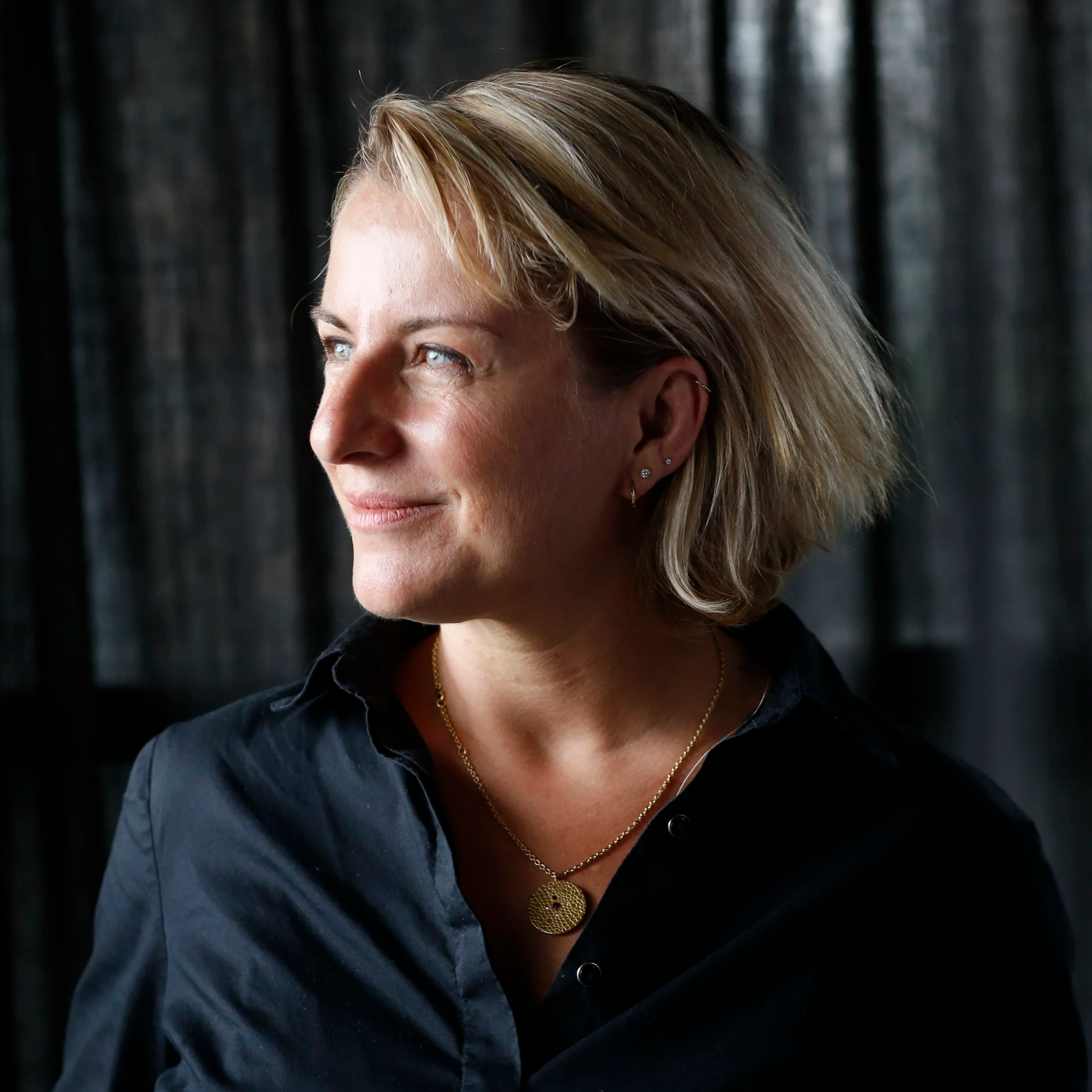 Close-up of a woman with blonde hair wearing multiple earrings, a gold necklace, and a dark shirt, looking to the side with a black curtain background.