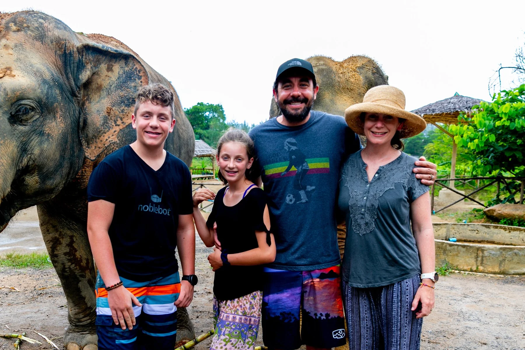A family of four posing with an elephant at a zoo or wildlife park. The adults and children are smiling and standing close together, with the elephant in the background. The setting appears outdoors with greenery and a fence.