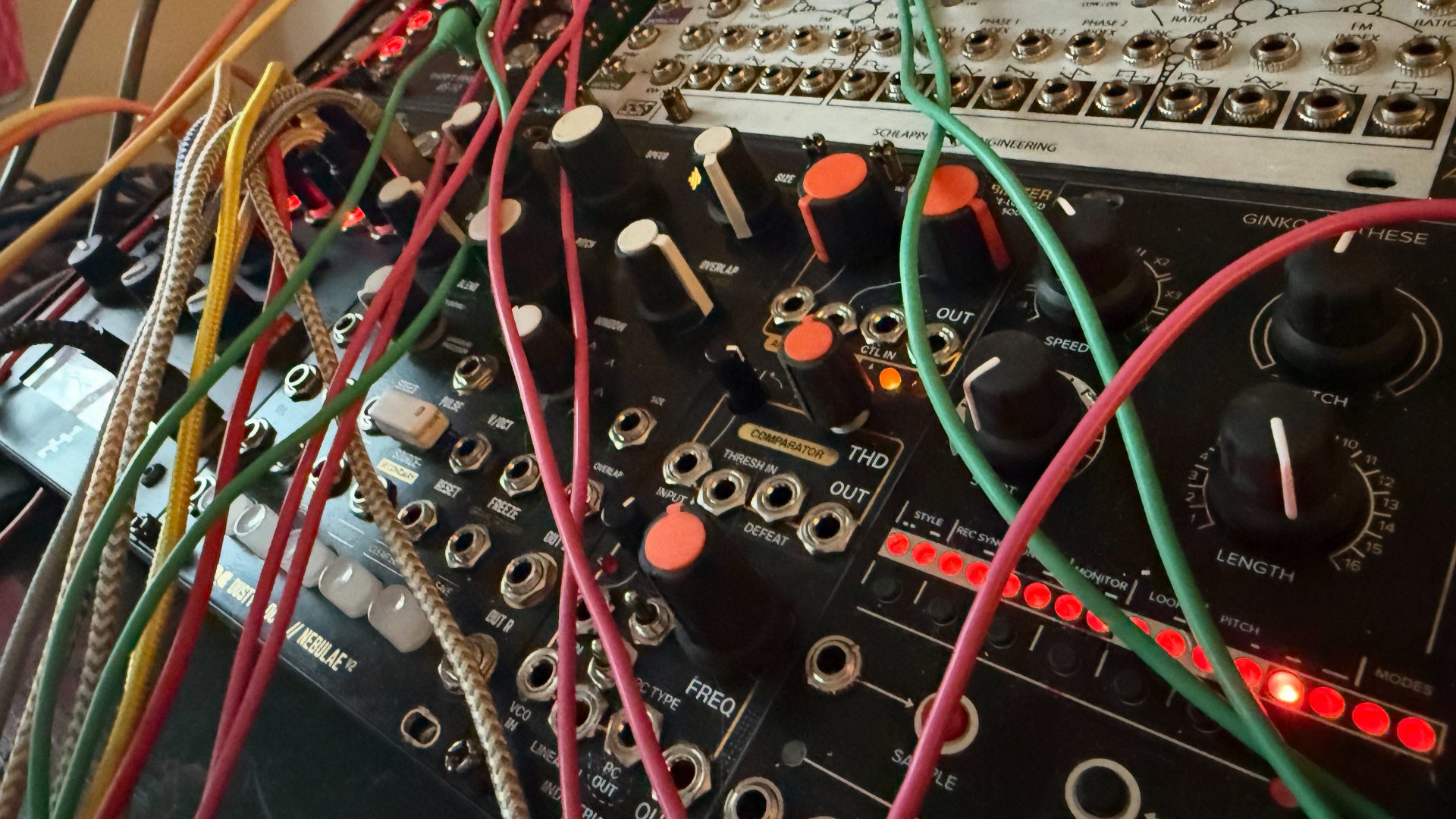 Close-up of a modular Eurorack music synthesizer with various colorful patch cables connected to different sliders, knobs, and input/output ports.