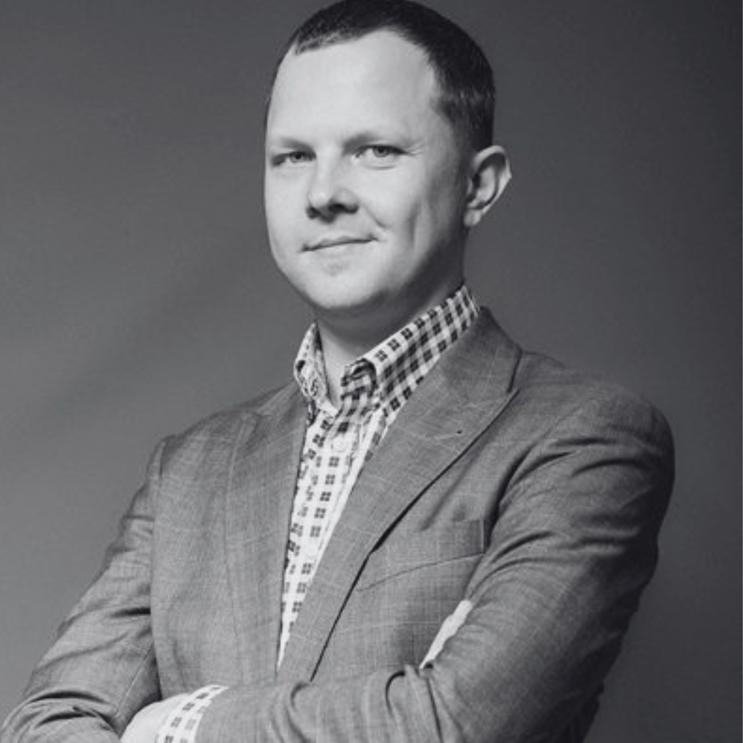 Black and white portrait of a man with slicked-back hair wearing a checkered shirt and a blazer.