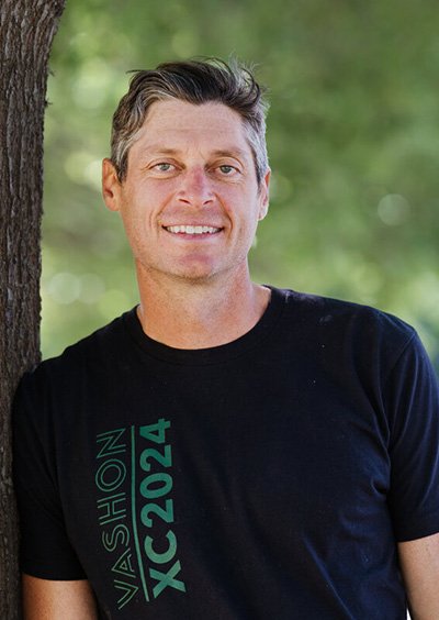 A smiling man standing outdoors near a tree with a blurred green background.