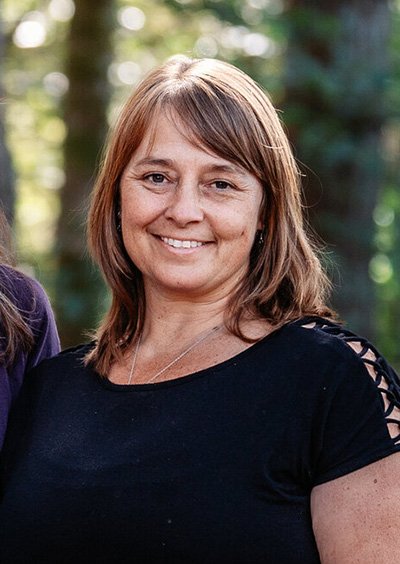 A woman with shoulder-length light brown hair, wearing a black top with lace-up detail on the sleeves, smiling outdoors with a blurred forest background.
