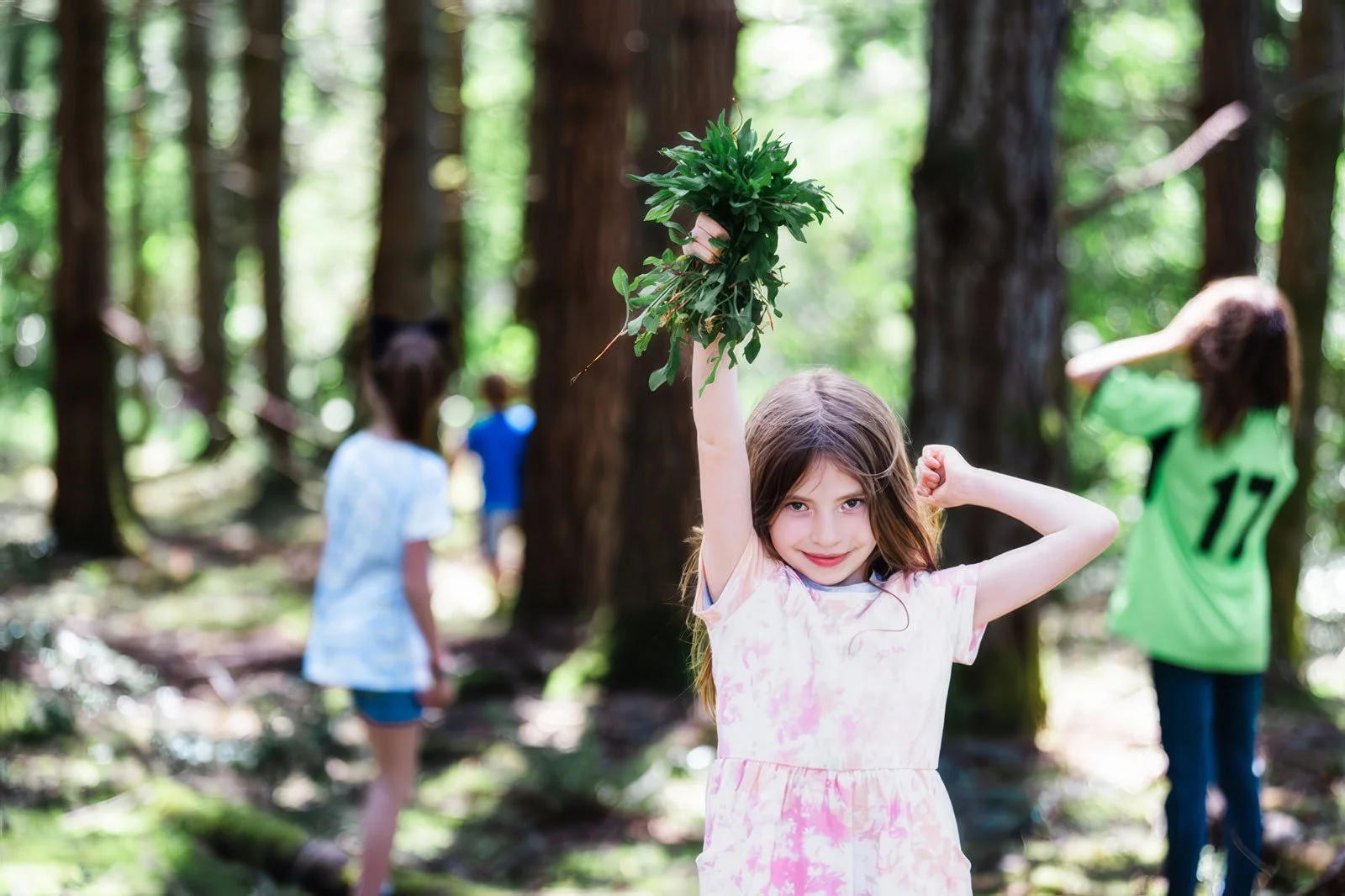 A young girl holding a bunch of green leaves high in the air with a confident expression in a forest, other children in the background.