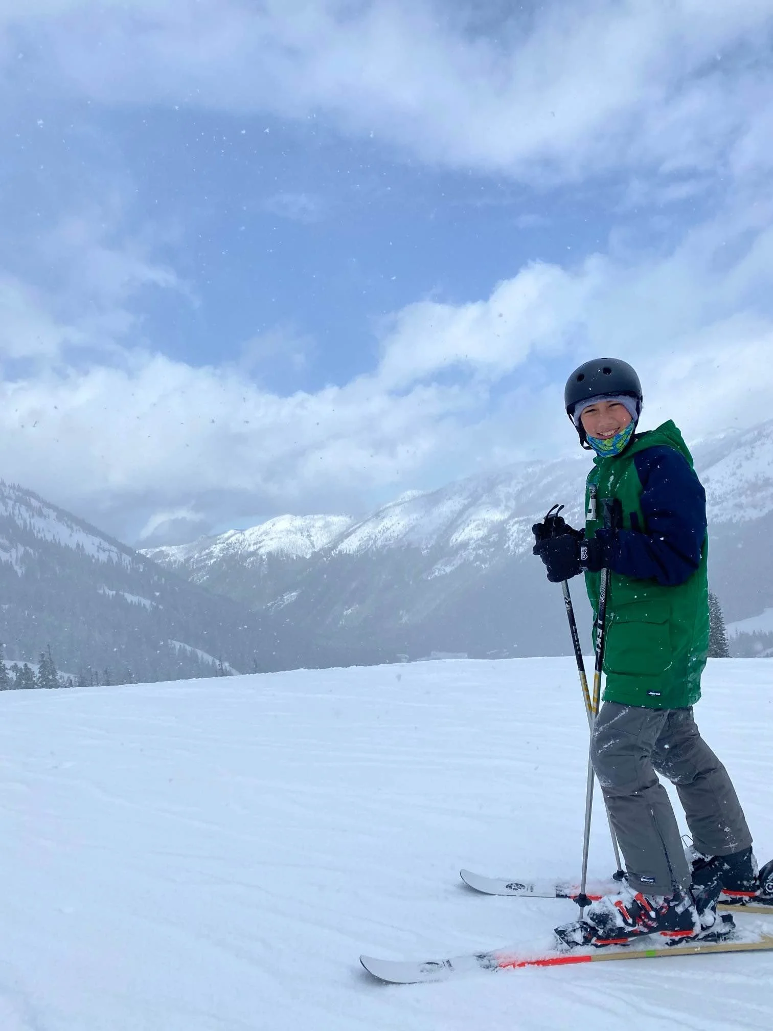 A young boy smiling and dressed in winter gear, holding ski poles, and standing on skis in a snowy mountain landscape with cloudy sky in the background.