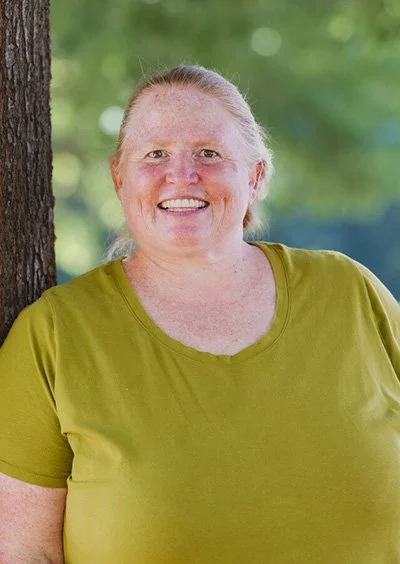 A smiling woman with light skin and blonde hair tied back, wearing a green t-shirt, standing outdoors near a tree with green foliage in the background.