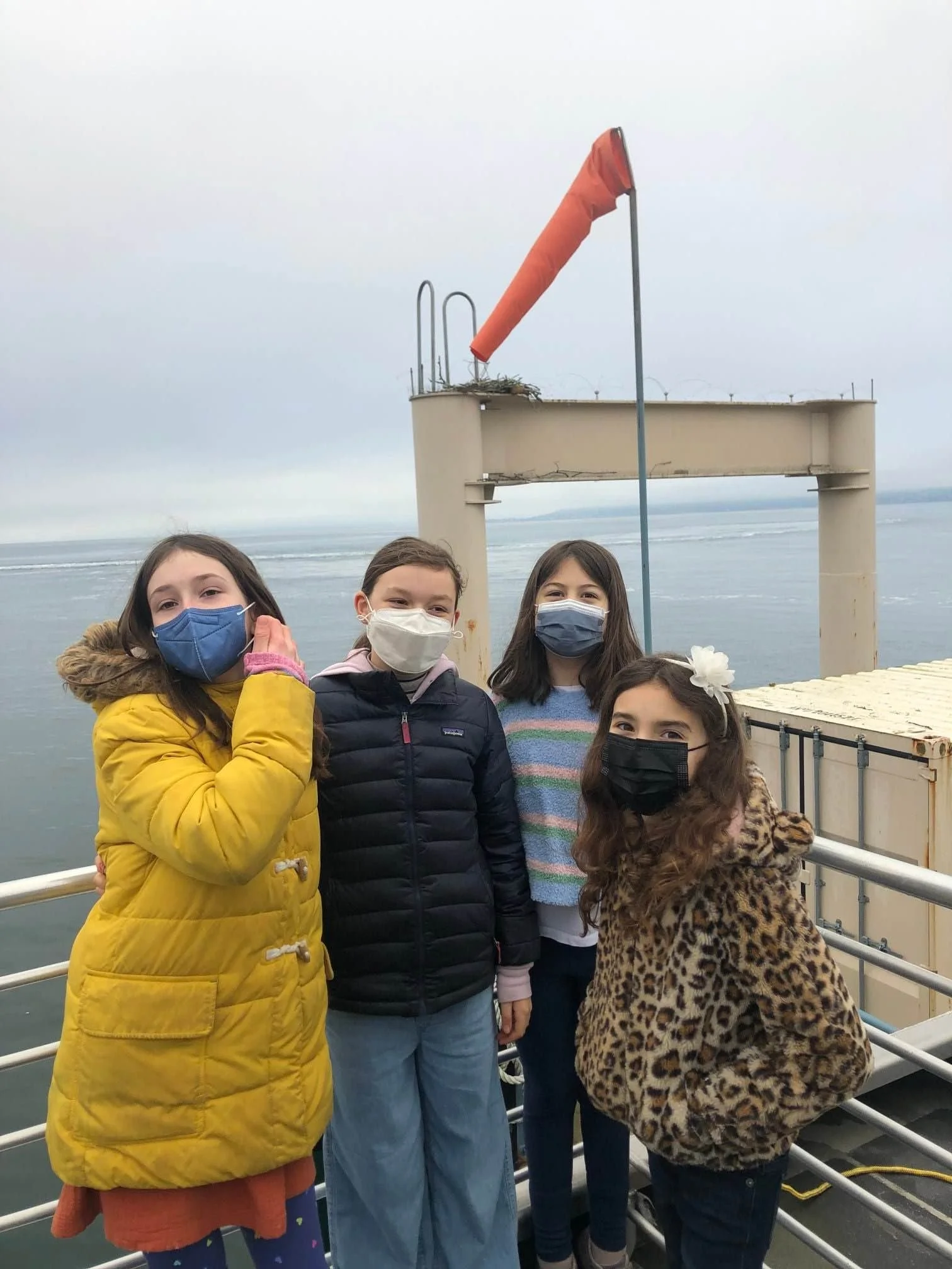 Four children wearing masks and warm clothing stand on a boat or ferry with water and a cloudy sky in the background. An orange windsock and a beige structure are visible behind them.