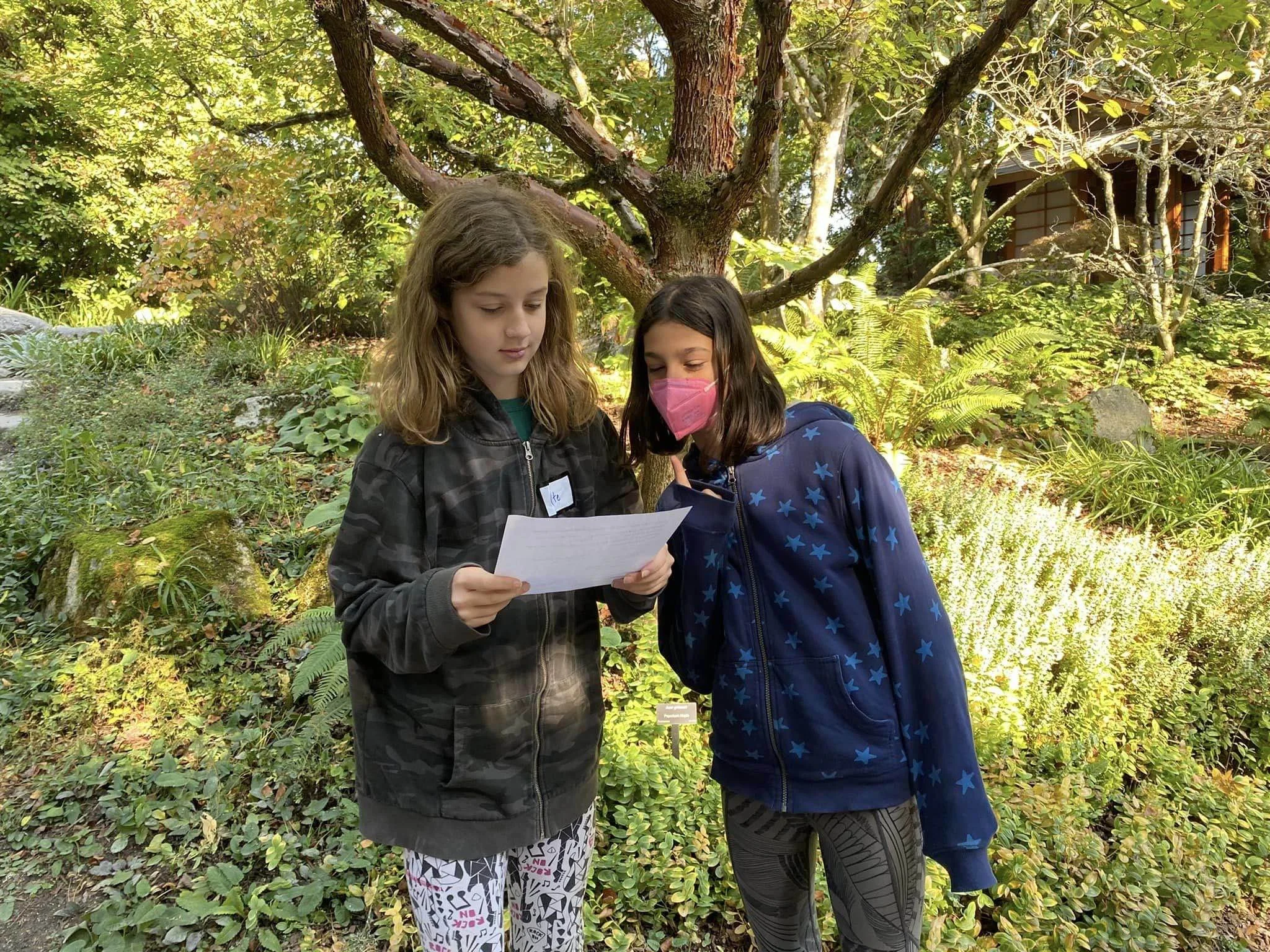 Two young girls standing outdoors in a garden with lush green foliage and a large tree, looking at a piece of paper together. One girl is wearing a pink face mask, and the other has long, wavy hair.