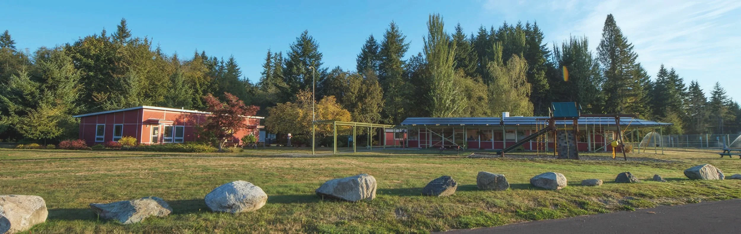 A schoolyard with a red building, playground equipment, rocks, and surrounding trees.
