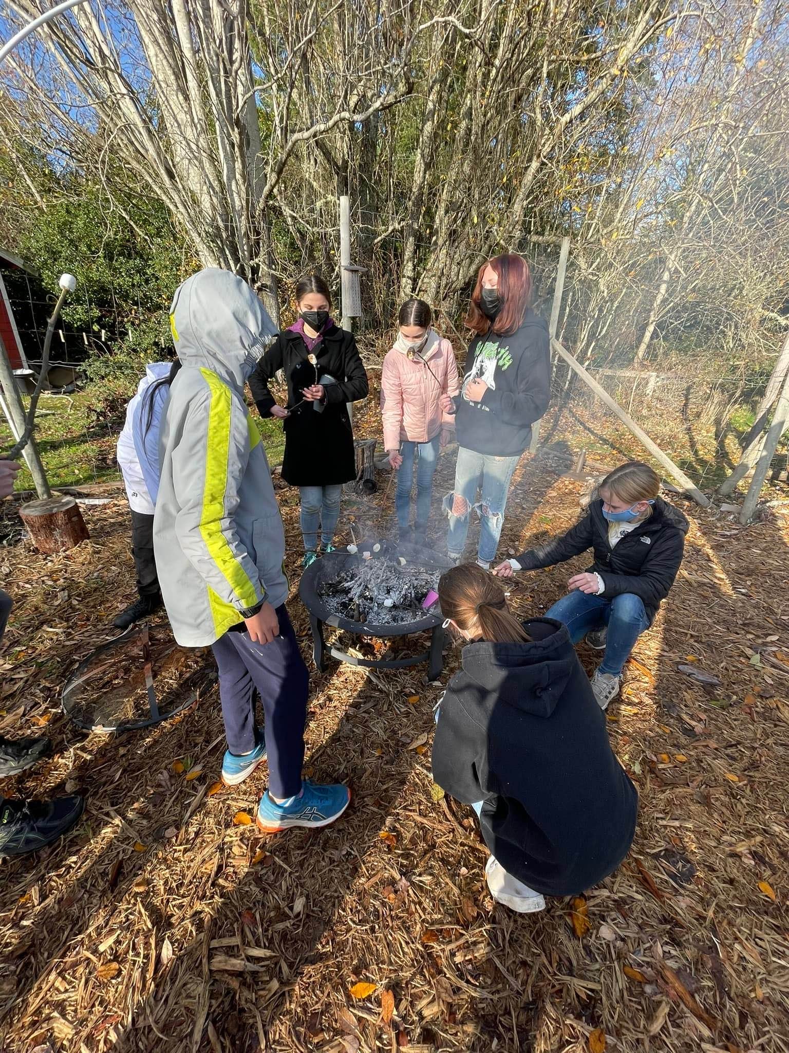 A group of people, wearing masks, gathered around a small outdoor fire in a wooded area during fall, with fallen leaves on the ground.