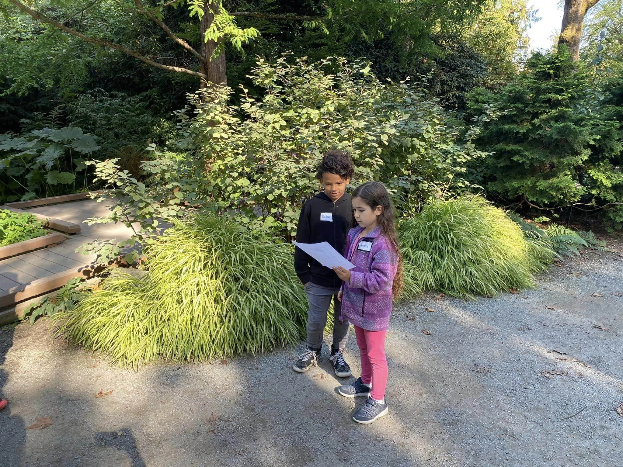 Two children, a boy and a girl, are standing outdoors on a gravel path surrounded by lush green plants and trees. The girl is reading from a piece of paper while the boy watches. The girl wears a pink and purple patterned jacket and pink pants, and t