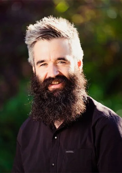 Man with a full beard and gray hair, wearing a black shirt, standing outdoors with blurred green foliage in the background.