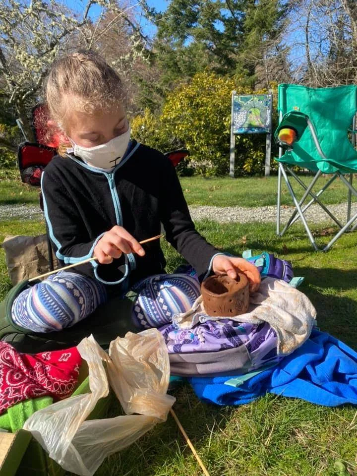A girl sitting outside on green grass, crafting pottery with clay. She is wearing a face mask, a black jacket with teal accents, and patterned leggings. There are camping chairs, bags, and outdoor objects around her, with trees and a blue sky in the 