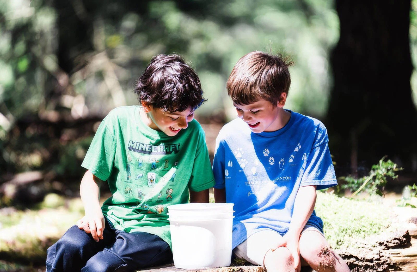 Two boys smiling and looking at a white bucket outside in a wooded area.
