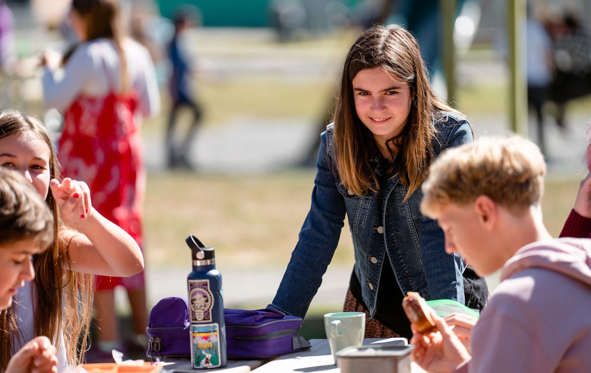 A group of young people gathered outdoors around a table during daytime, engaging in conversation and activities, with a focus on a girl with long brown hair leaning on the table.