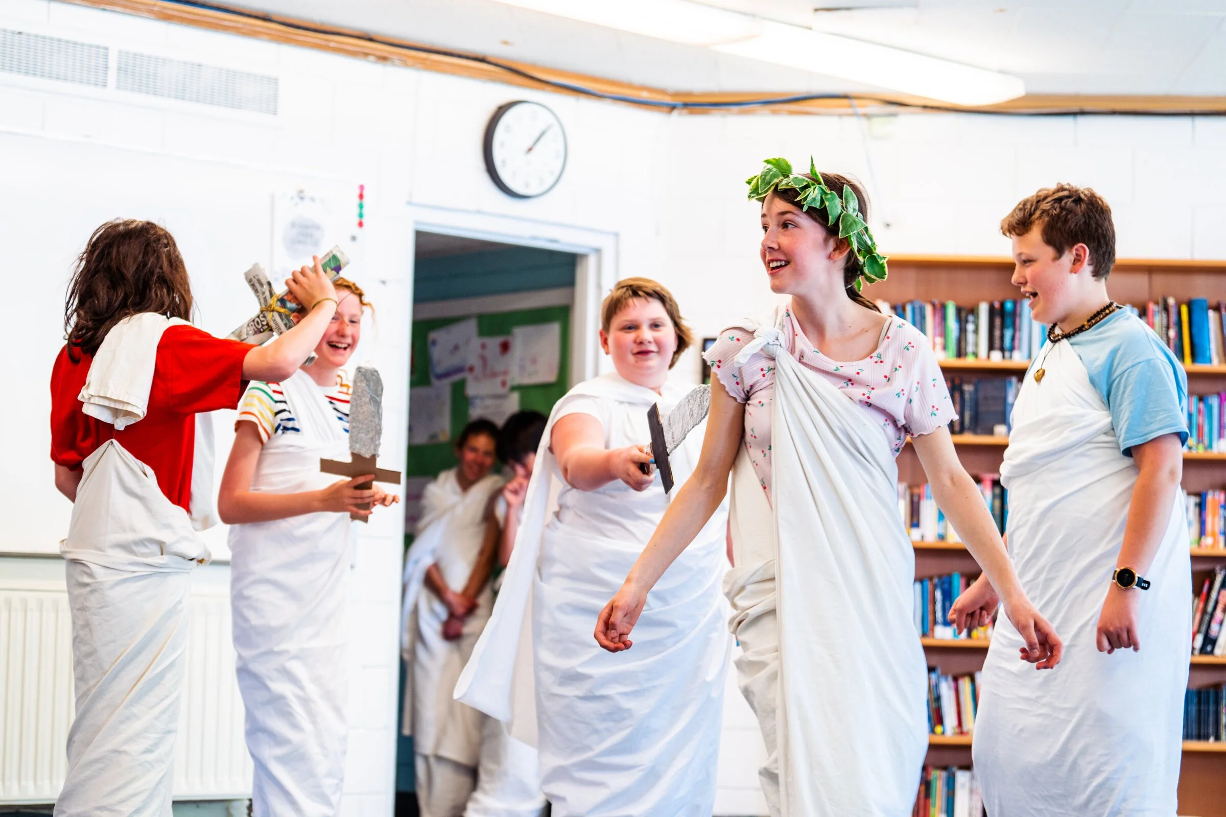 Children dressed in costume for a play, participating in a Greek or Roman themed performance in a school library.