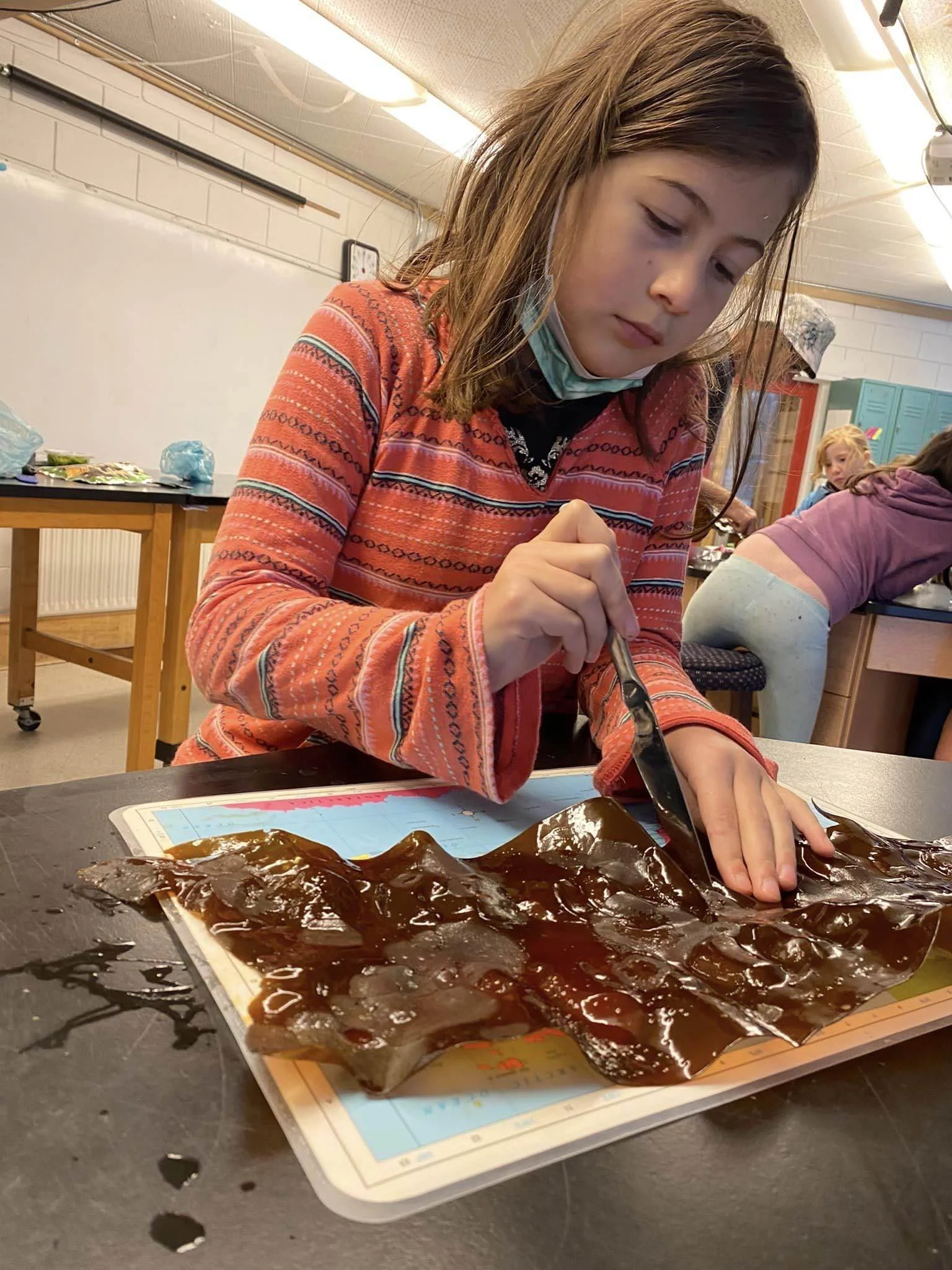 A girl is working on an art project involving painting or coloring on a sheet of paper. There are multiple sheets of paper and dark paint or ink on a tray in front of her. The setting appears to be a classroom or art studio.
