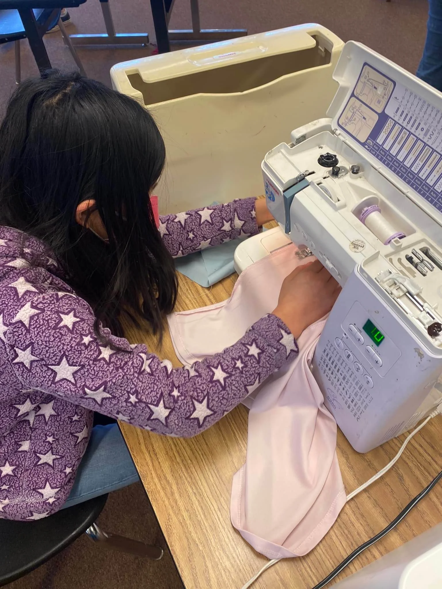 A young girl is sewing on a white sewing machine at a wooden table, wearing a purple fleece jacket with star patterns, in a room with chairs and tables.