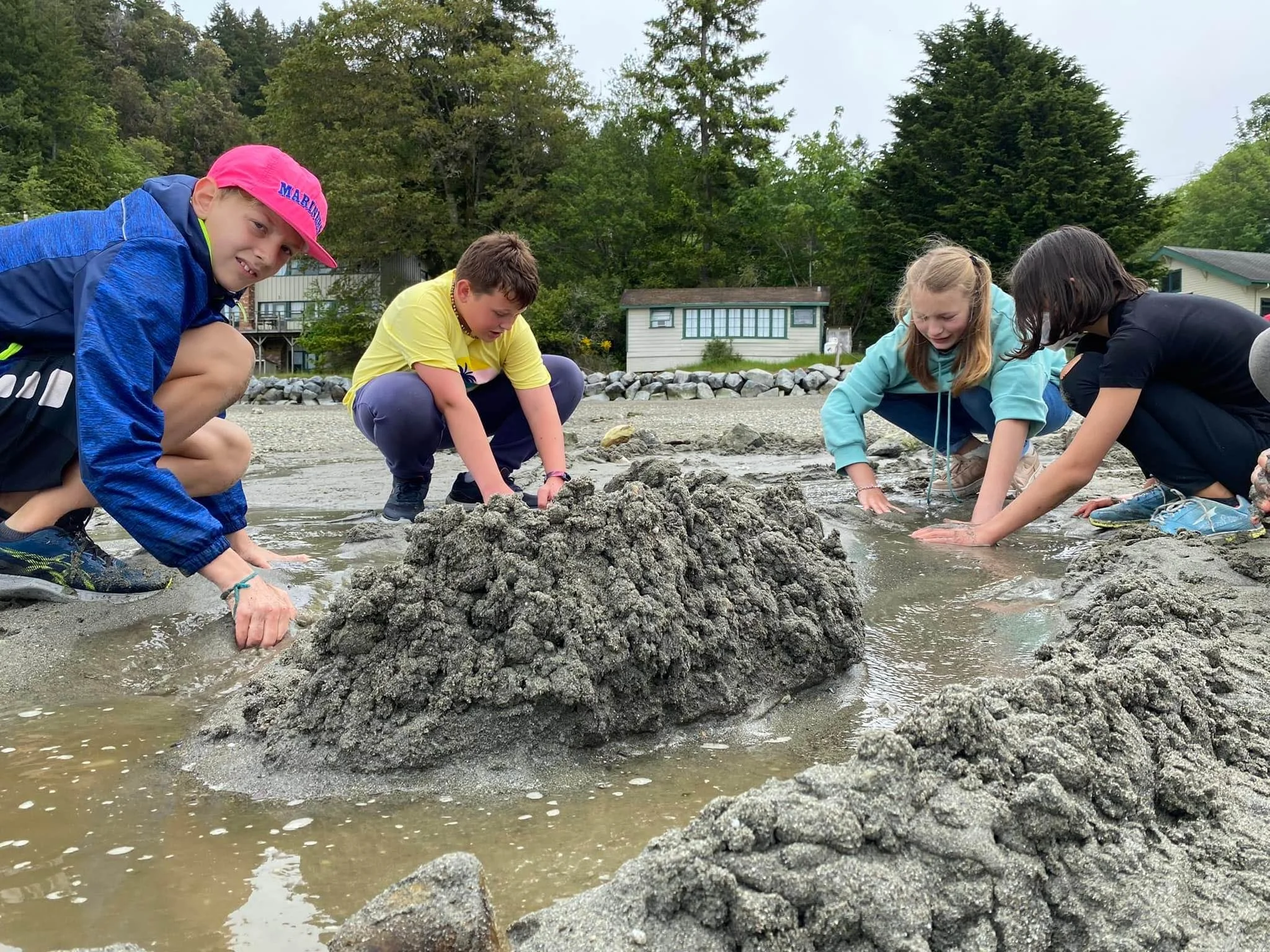 Four children are kneeling on wet sand near the water's edge, examining the shoreline with their hands in the sand. They are outdoors, with trees and small houses in the background, under an overcast sky.