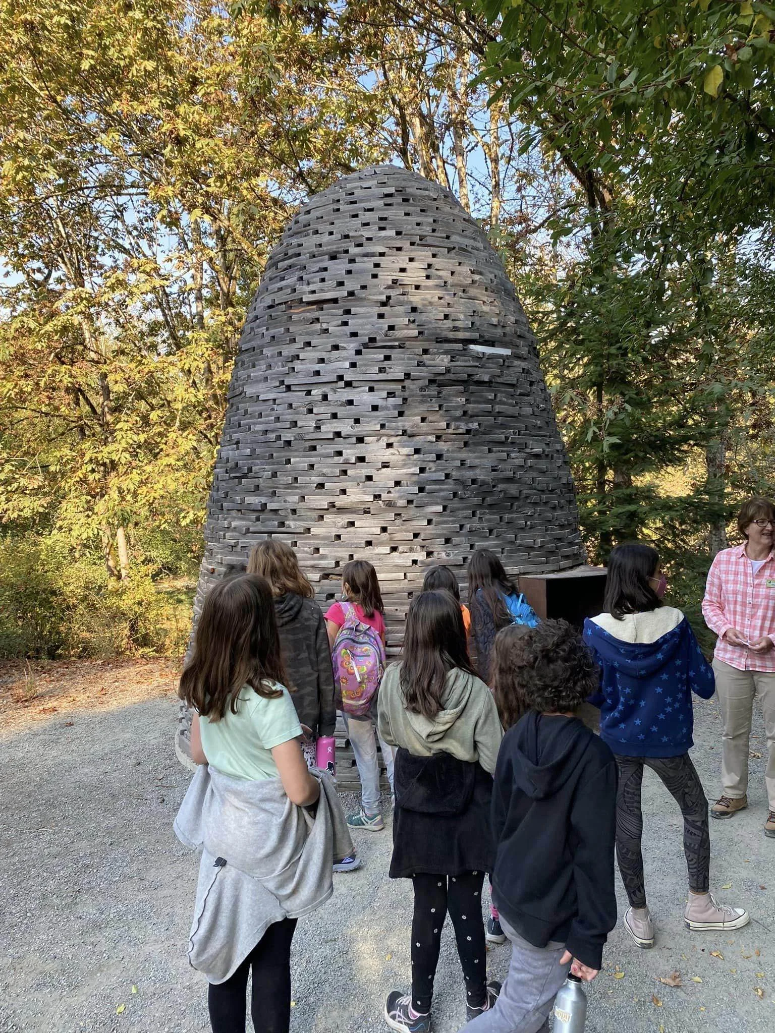 A group of children and an adult in a park, standing around a large wooden bee hive sculpture, surrounded by trees with autumn leaves.
