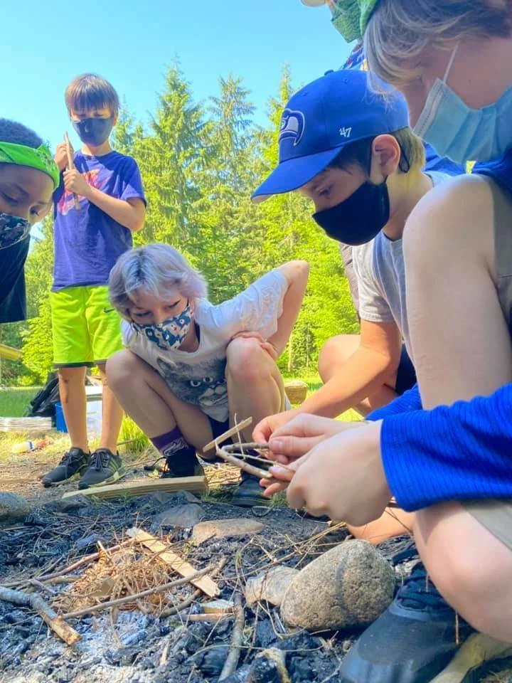 Children gathered around a campfire outdoors, roasting marshmallows on sticks, with trees and a blue sky in the background.