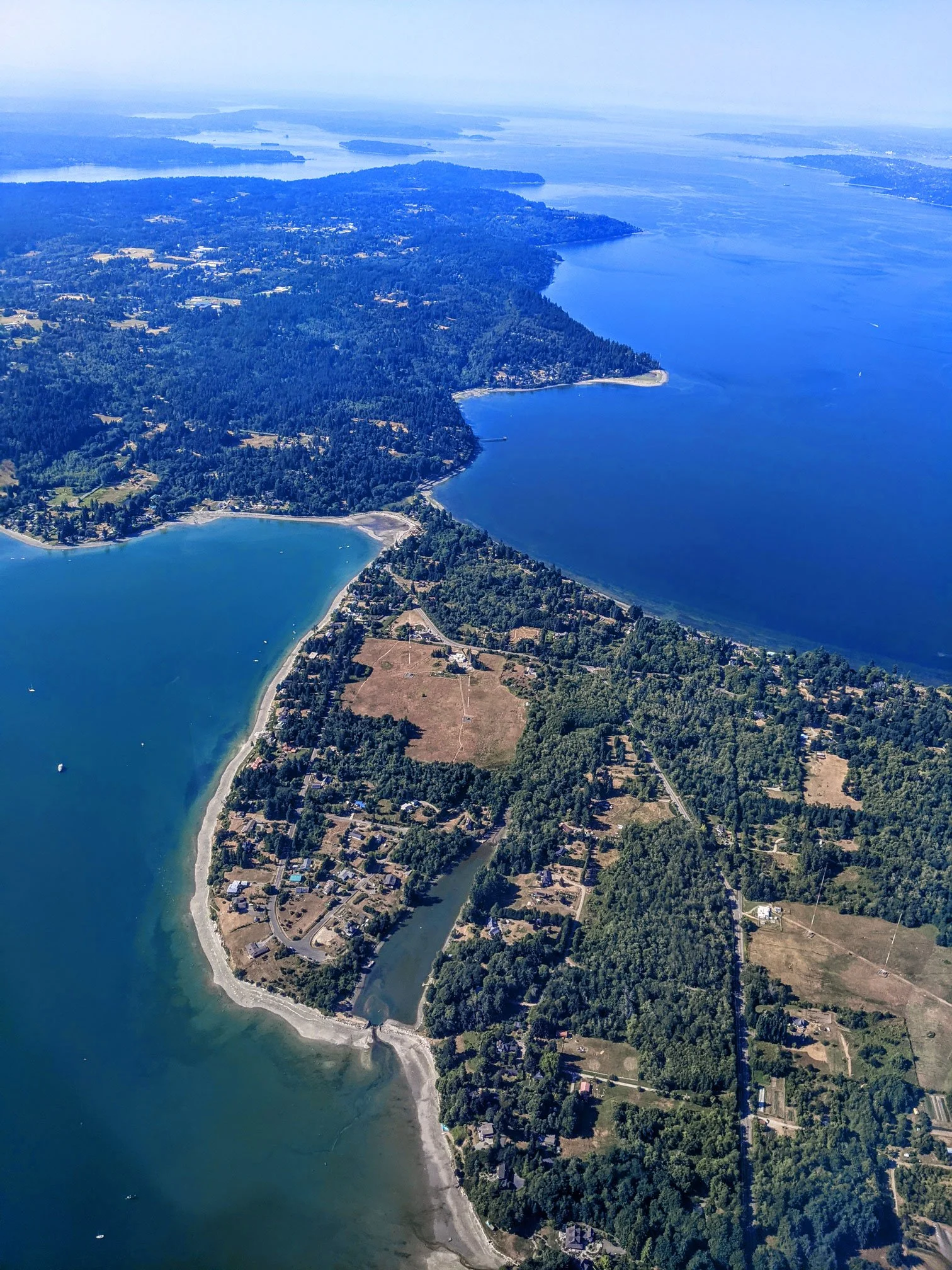 Aerial view of a large coastal area with a mix of forested land, residential homes, and bodies of water including a large bay, a river, and several islands in the distance.