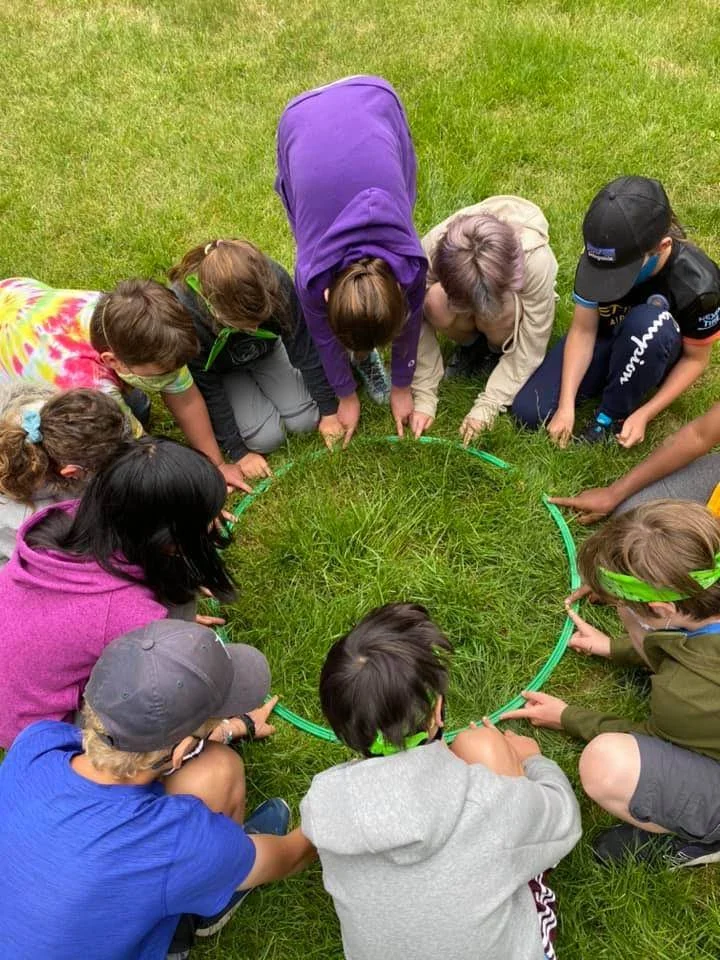 Group of children and two adults gathered around a small patch of grass, using a green plastic hoop, possibly for an outdoor activity or game.