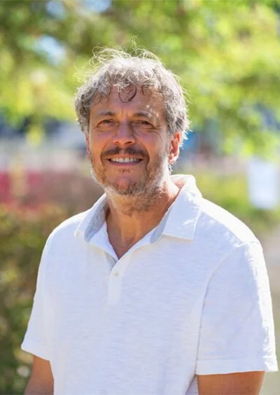 A man with gray curly hair and a beard smiling outdoors, wearing a white polo shirt, with blurred green trees in the background.