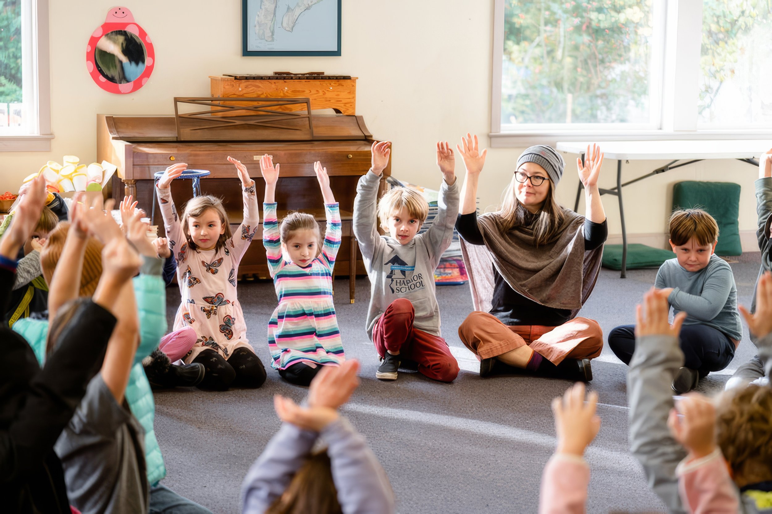 A classroom scene with children sitting cross-legged on the floor, raising their hands, and a woman with glasses and a gray beanie leading them in an activity. The room has a piano, a mirror with a strawberry character decoration, and windows with natural light.