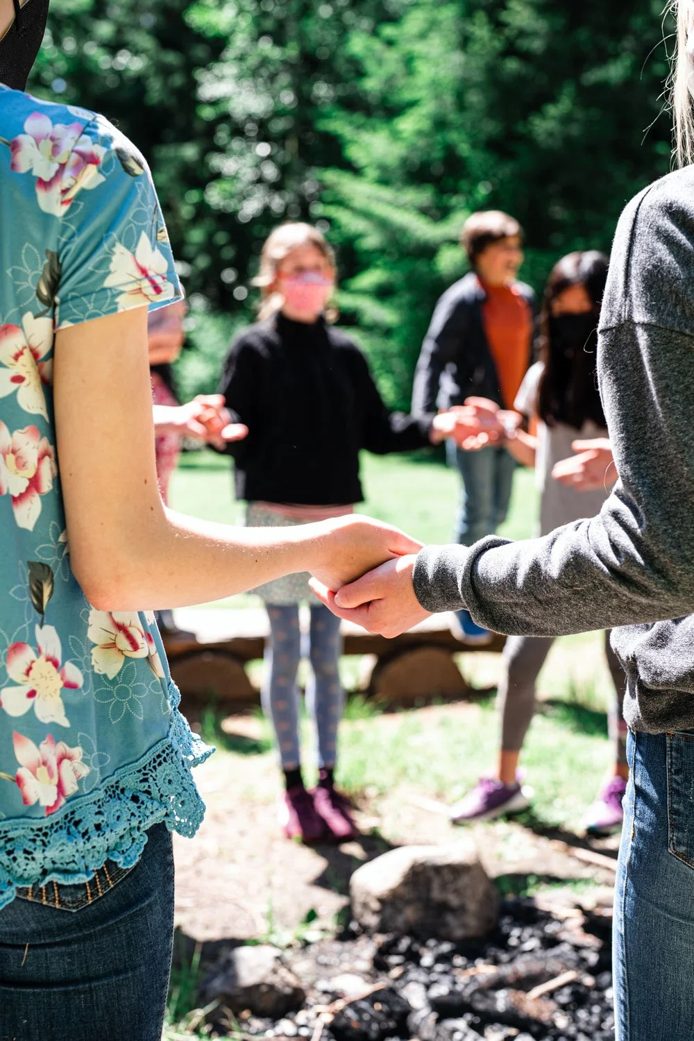 Two people holding hands in the foreground, with a group of people in the background outdoors on a sunny day, surrounded by trees.