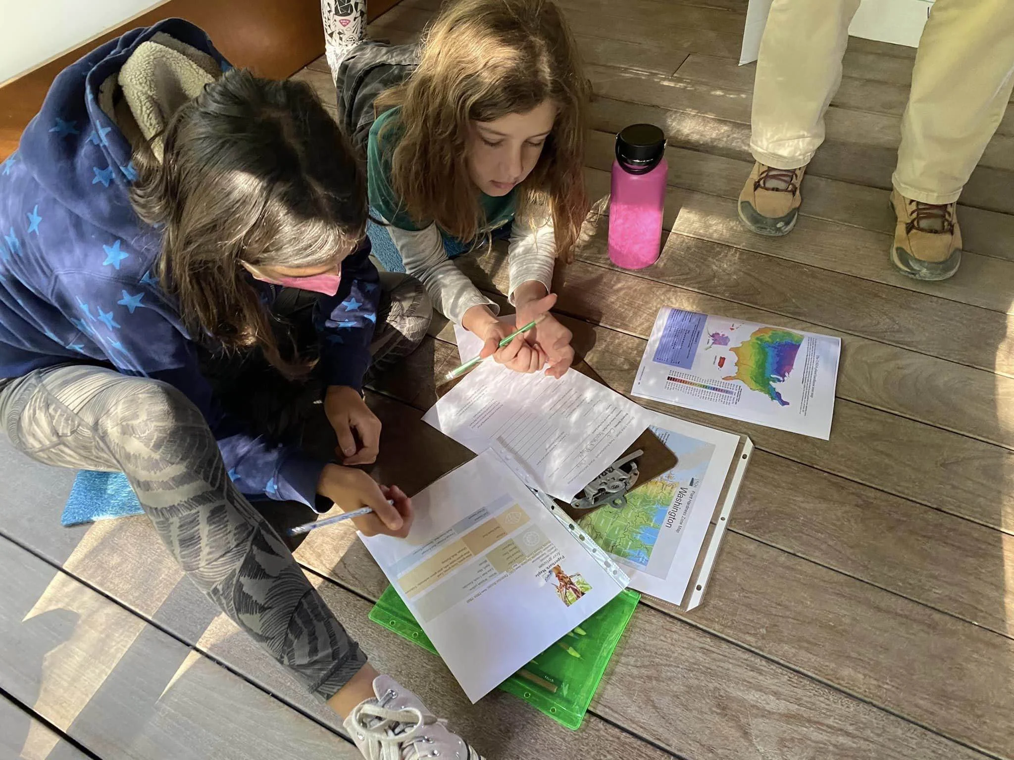 Two young women sitting on the wooden floor, surrounded by maps, documents, and a pink water bottle, engaged in a discussion or studying. One is holding a pencil,
while the other looks at the materials. A third person stands nearby, only showing thei
