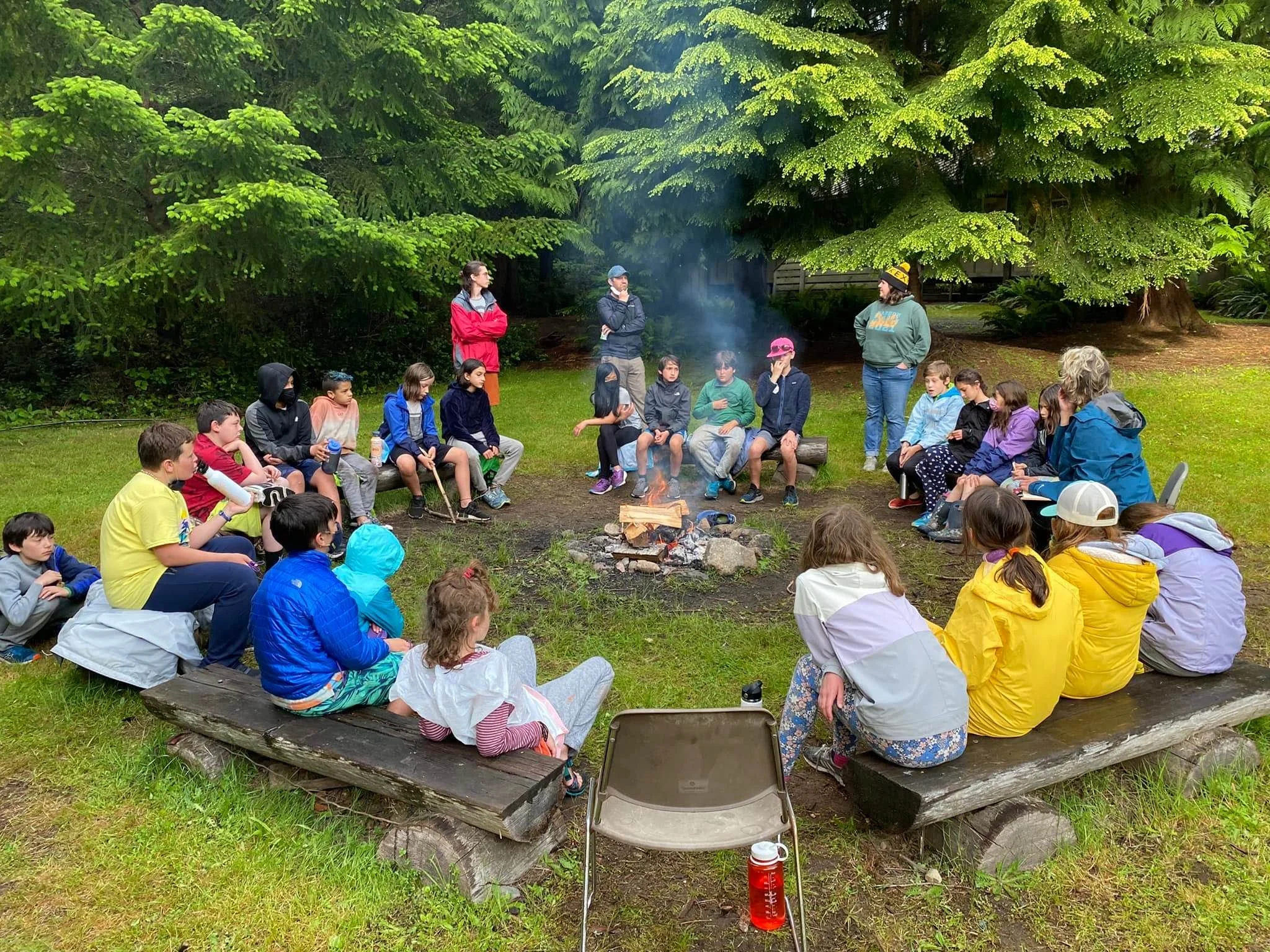A group of children and a few adults sitting and standing around a campfire in a wooded area with large green trees.