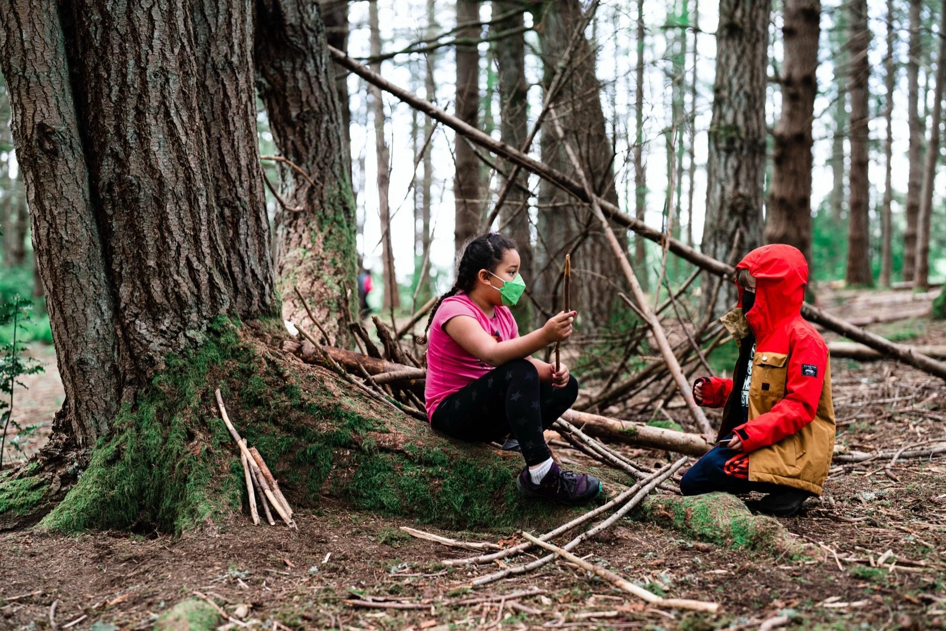 Two children, a girl and a boy, in a forest. The girl is sitting on the ground near a large tree, wearing a pink shirt and a green mask, holding a stick. The boy is kneeling near the girl, wearing a red jacket with a hood and a black mask. They are s