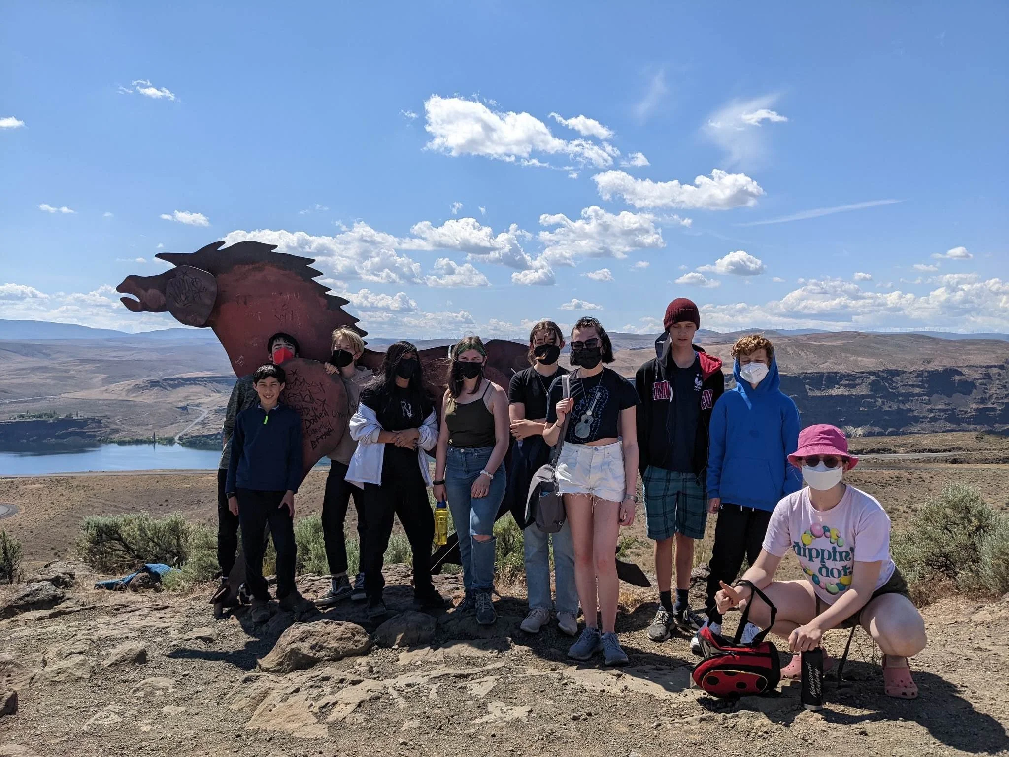 Group of children and teenagers standing outdoors on rocky terrain, with a large red sculpture of a dragon's head behind them, in a scenic landscape with mountains, a lake, and a blue sky with scattered clouds.