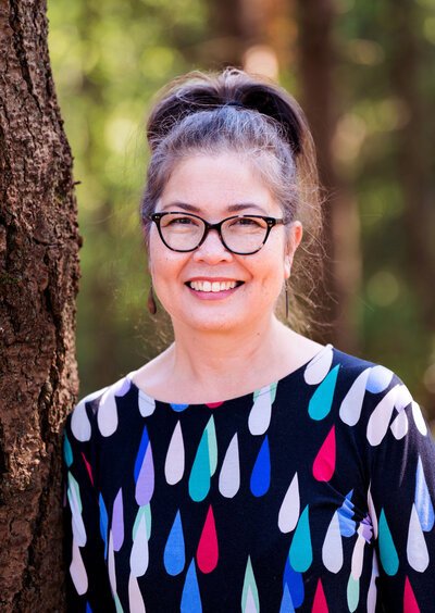 A woman with gray hair tied back in a high ponytail, wearing black glasses and a colorful teardrop patterned top, standing outdoors next to a tree in a forested area.