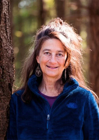 A woman with long wavy hair smiling outdoors in a forest, wearing a blue fleece jacket and earrings.