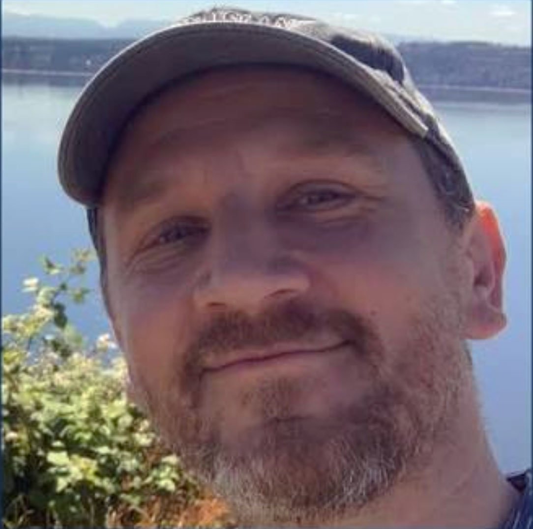 Close-up of a man outdoors near water, wearing a cap, with a beard and mustache, smiling.