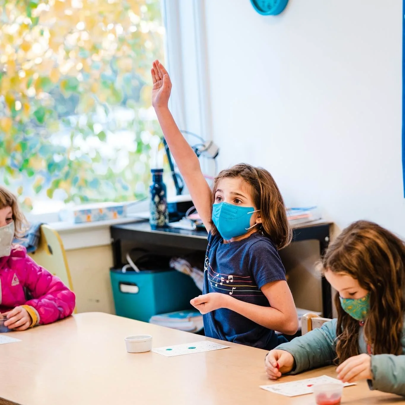 A classroom scene with children wearing face masks, a girl in the middle raising her hand, and other children seated at a table, engaged in activity.